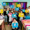 Children in an art class hold up their blue and yellow drawings of birds, smiling in front of a colorful wall with lights.
