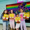 Four girls hold up yellow cartoon paintings with big eyes and a red tie, posing in a colorful art studio with a neon “Honest Art” sign.