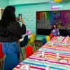 Girl in a purple sweater poses for a photo in a bright rainbow party room with a striped table and neon art on the wall.