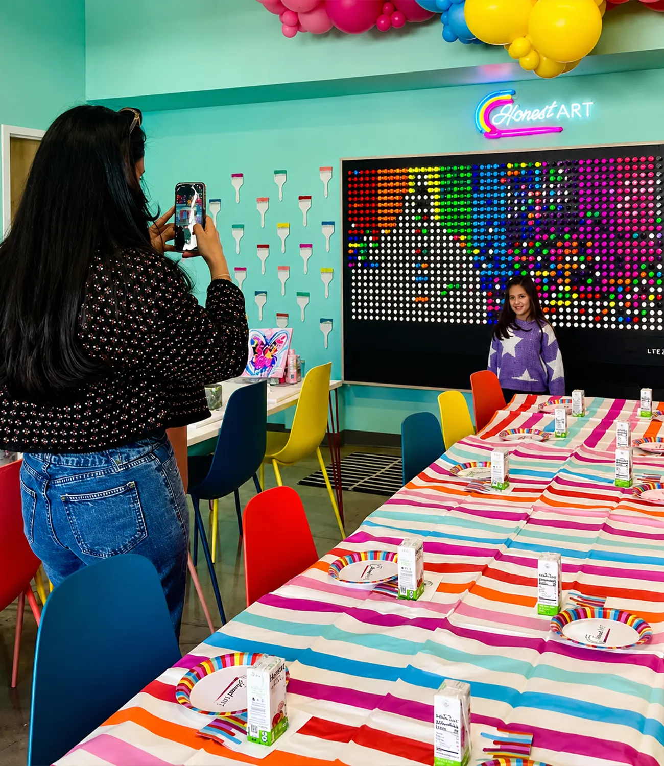 honest-art-web-4 | LiteZilla Girl in a purple sweater poses for a photo in a bright rainbow party room with a striped table and neon art on the wall.