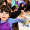 Child with glasses and a purple apron paints a colorful clay figure at a white table in a bright art classroom, with classmates and a rainbow wall in the background.