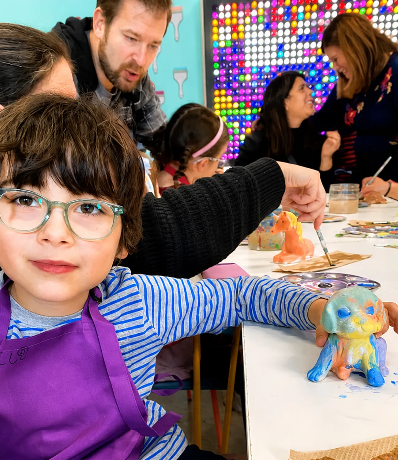 honest-art-web-5 | LiteZilla Child with glasses and a purple apron paints a colorful clay figure at a white table in a bright art classroom, with classmates and a rainbow wall in the background.