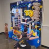 Kids interact with a large colorful hands-on science wall featuring tubes, gears, and levers.