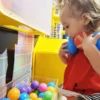 Toddler playing at a ball arcade game, holding a blue and an orange ball up to her neck in a bright, colorful setting.