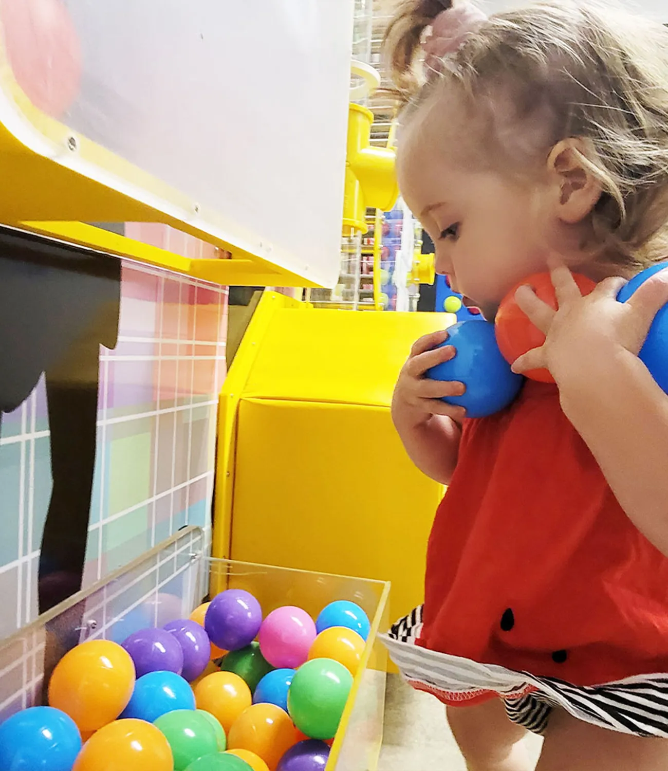 ball-launcher-10 | LiteZilla Toddler playing at a ball arcade game, holding a blue and an orange ball up to her neck in a bright, colorful setting.