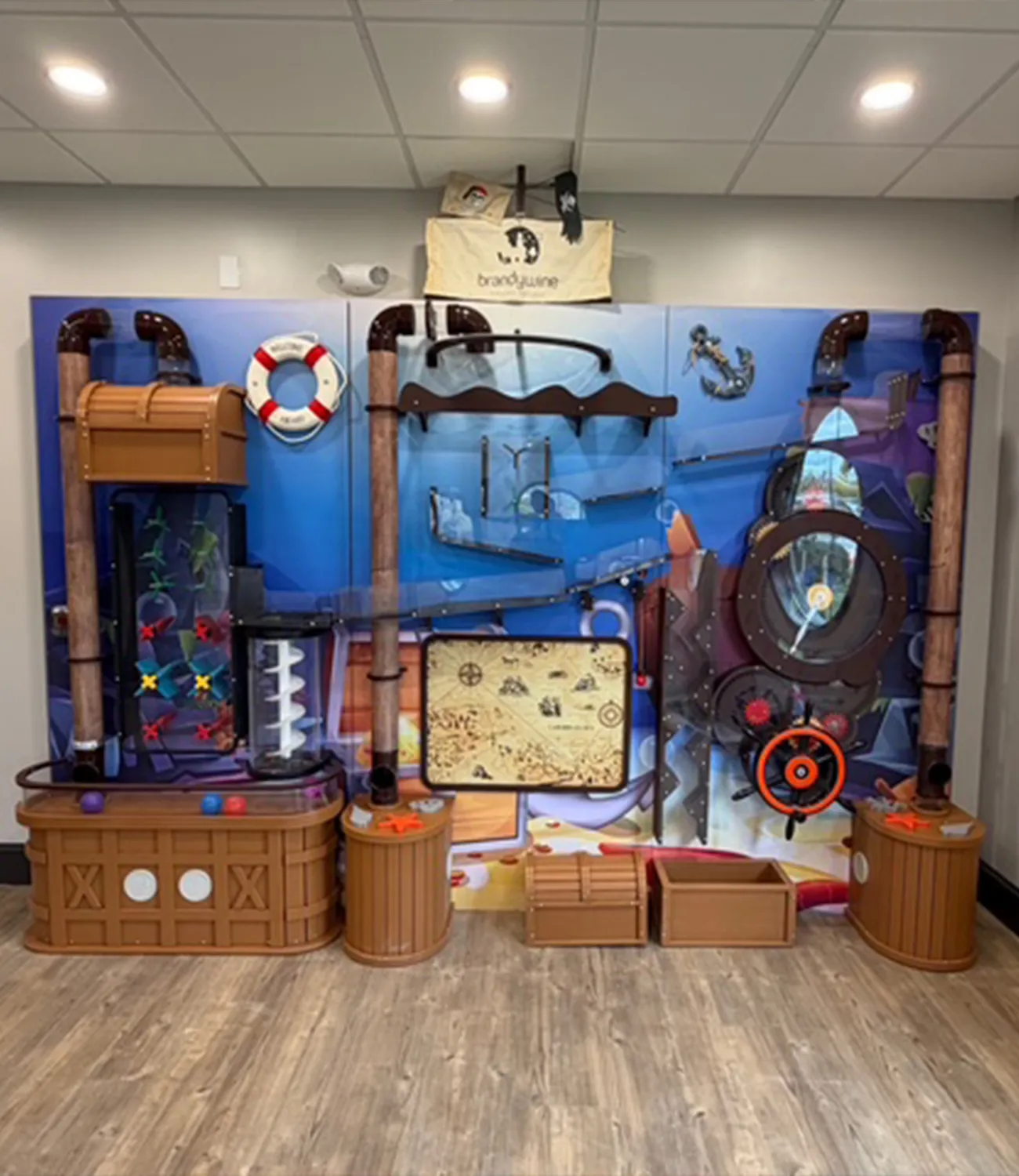 ball-launcher-3 | LiteZilla Colorful pirate/treasure-themed indoor play wall for kids, with tubes, a life preserver, a steering wheel, and treasure chests set against a blue mural.