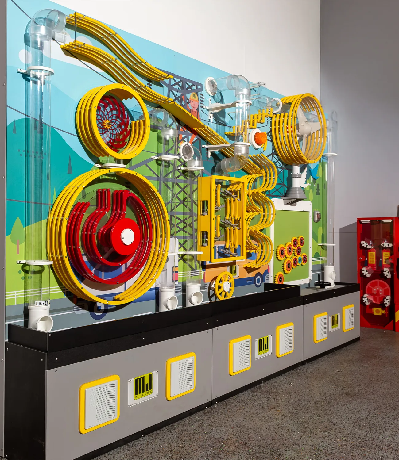 ball-launcher-5 | LiteZilla Colorful kinetic mechanical sculpture with yellow circular gears and winding tubes mounted on a mural-filled panel in a museum setting.
