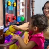 Child in a pink dress reaches for a yellow ball in a colorful arcade ball-toss game, with an adult watching nearby.