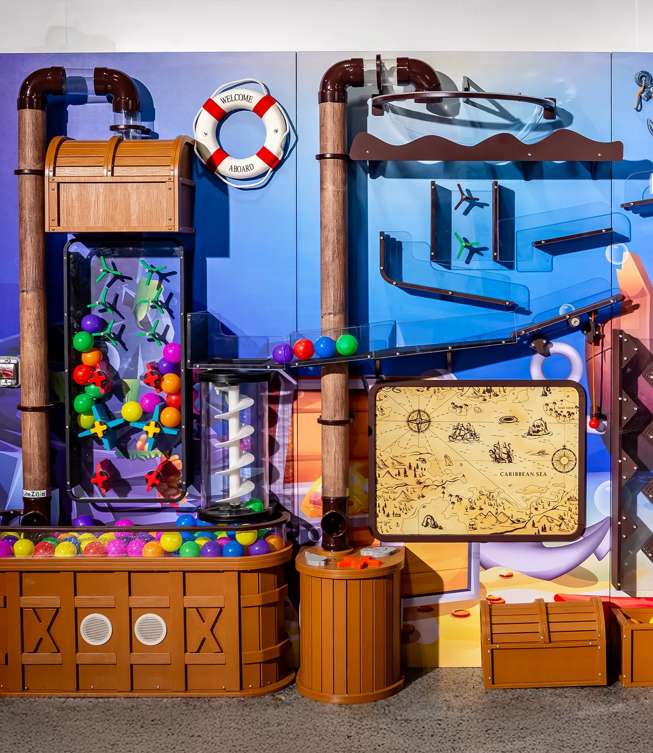 ball-launcher-7 | LiteZilla Colorful indoor playground wall featuring brown barrels, a spiral corkscrew, a glass ball maze, and a pirate-map panel on a blue background.