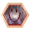 Hexagonal, sequin-covered badge with a red smiling face on a pale wooden frame.